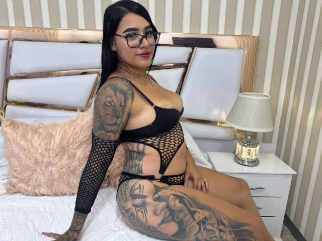 MelodyRuso mit Tattoos, schwarzer Netzstrumpfhose, Brille und pinkem Lippenstift, sitzt auf dem Bett, zeigt ihre Brüste und ihren Hintern, mit einem flauschigen pinken Kissen und einem weißen Nachttisch..