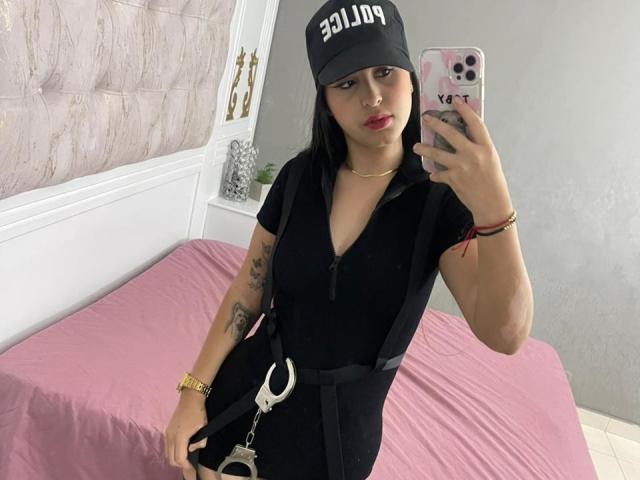 ZoeCastelli with black cap, red lipstick, black zip-up dress, handcuffs, tattoo on left arm, taking selfie in bedroom, pink bed, gray wall, gold jewelry, and pink phone case.