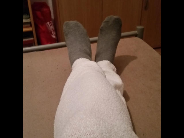 Le gambe di CashLadyVivian-hot, con calze grigie e un asciugamano bianco, sdraiate su un tappeto beige, con una porta di legno sullo sfondo, texture morbida e confortevole, atmosfera rilassata e accogliente..