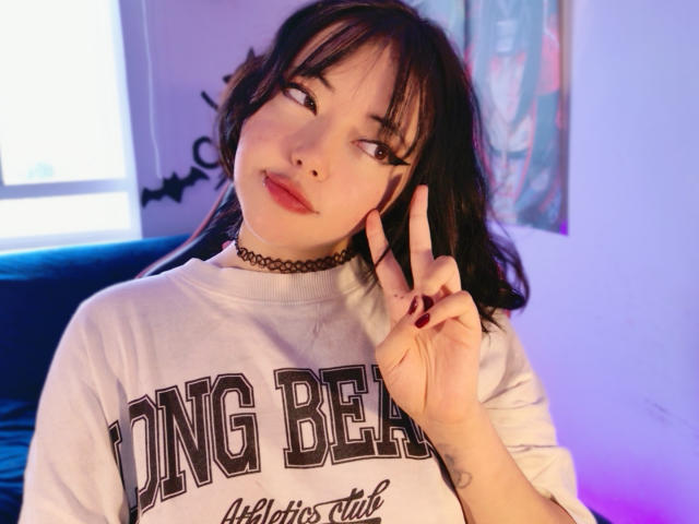 Arieyaara with dark hair, bangs, and red lipstick, wearing a white shirt, black choker, and red nails, flashing a peace sign with her right hand.