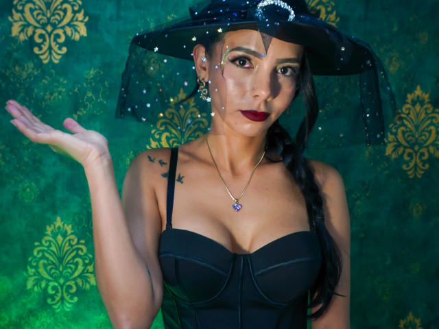 EvelinLuna with dark hair in a braid, wearing a black dress, sheer hat with stars, red lipstick, necklace, raising her hand, showing her tits, green background.