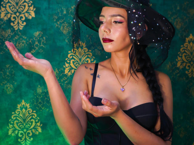EvelinLuna with dark red lipstick, black strapless dress, braid, black witch hat with stars, holding open hands, small tits, green and gold background, tattoo on right shoulder.