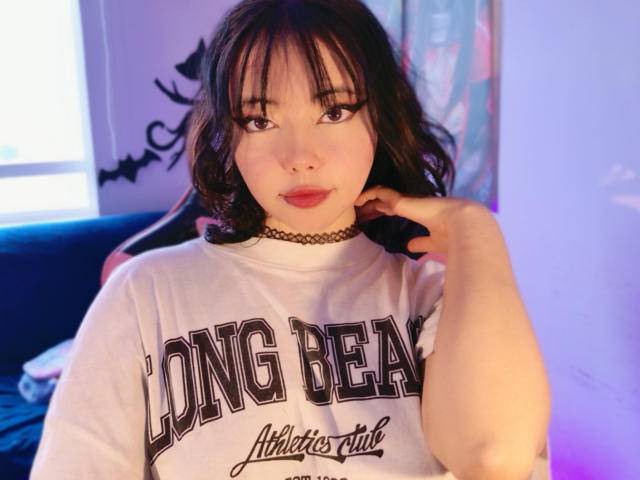 Arieyaara, avec ses cheveux bruns mi-longs, porte une chemise blanche avec l'inscription "Long Beach", un collier noir et touche son cou. Elle a les lèvres roses et une expression légèrement surprise..