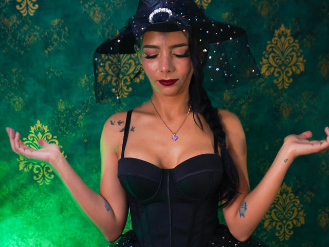 EvelinLuna with dark makeup, wearing a black witch hat and corset, has her hands up, showing off tattoos on her arms and chest, with small tits and a cute face.