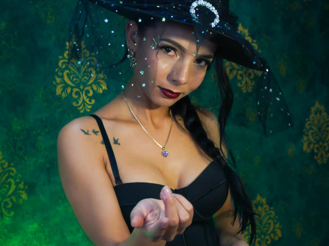 EvelinLuna, witch, wearing black dress, holding point, showing tits, braided black hair, dark lipstick, sparkly hat, gold necklace, bird tattoo on left shoulder, green background.