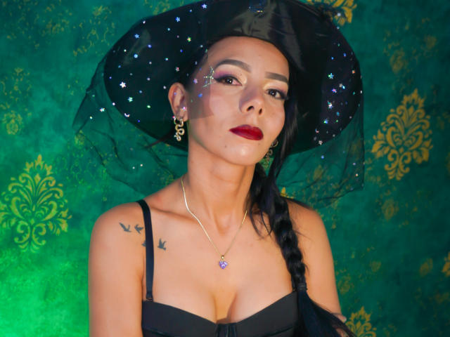 EvelinLuna with black witch hat, sparkly veil, braided hair, red lipstick, gold earrings, necklace, cleavage, small bird tattoos, green background, wearing black strap dress, looking confident.