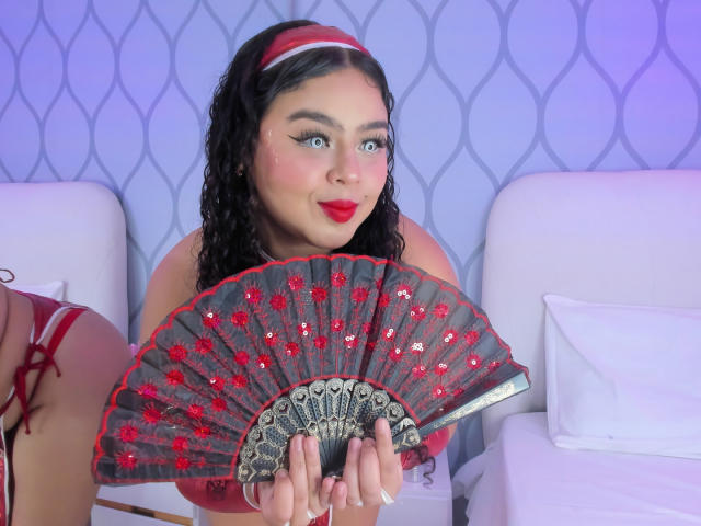 AprielKeith with curly black hair, red headband, red lipstick, holding a red and grey fan with red sequins, sitting between white pillows, showing off her cute face and tits.
