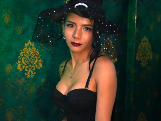 EvelinLuna with dark red lipstick, black strapless dress, and black witch hat with sparkles, showing her tits, green and gold background, looking at camera with a slight smile.
