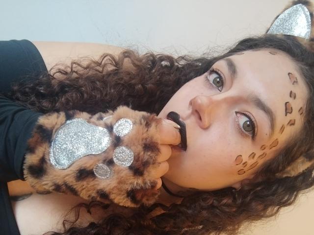 RubbyStrongX has curly brown hair, green eyes, and wears cat ears with glittery silver tips. She has leopard-print markings on her cheeks and wears black lipstick. Her right hand is covered in a furry, leopard-patterned glove with glittery silver paw pads. She's looking to the side with a slightly curious expression. The image is a close-up. RubbyStrongX's face is centered, and the focus is on her facial features and hand. Slang: "feline vibes."