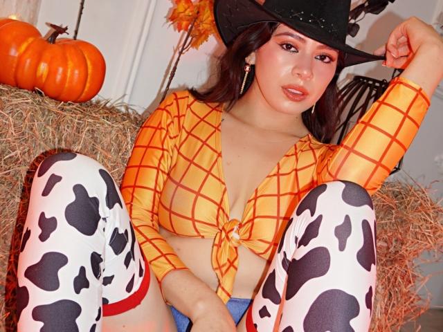 LindaMancini with large tits, wearing a plaid yellow top, black cowboy hat, cow-patterned thigh-highs, and blue shorts, sitting on hay with a pumpkin in the background.