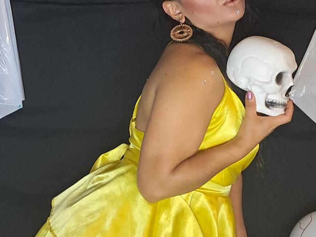 SamarAmour in a shiny yellow dress, holding a skull, with earrings and sparkling skin, her tits and ass partly visible, dark hair, black background.