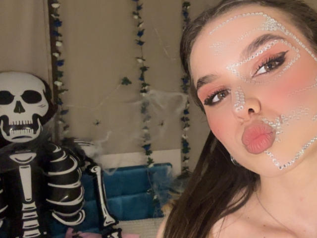 EmelyHailey with white glitter on her face, pouty lips, and bold eyeliner, next to a skeleton mask on the left. Her brown hair is down, and she's topless.