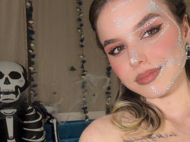 EmelyHailey has glitter on her face, big brown eyes with black eyeliner, pinkish cheeks, and red lipstick. She's next to a skeleton prop with a blue and white curtain.