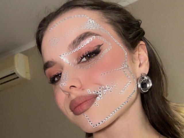 O rosto de EmelyHailey está coberto de bolhas de sabão brancas, com um blush rosa nas bochechas, e ela está usando um delineador preto grosso e brincos de argola prateados, olhando para a câmera..