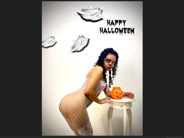 Macaroux with curly black hair wearing a white fishnet dress, bending over a white stool, holding an orange pumpkin. Boobs, ass, fishnet dress, pumpkin. Macaroux's tits are visible.