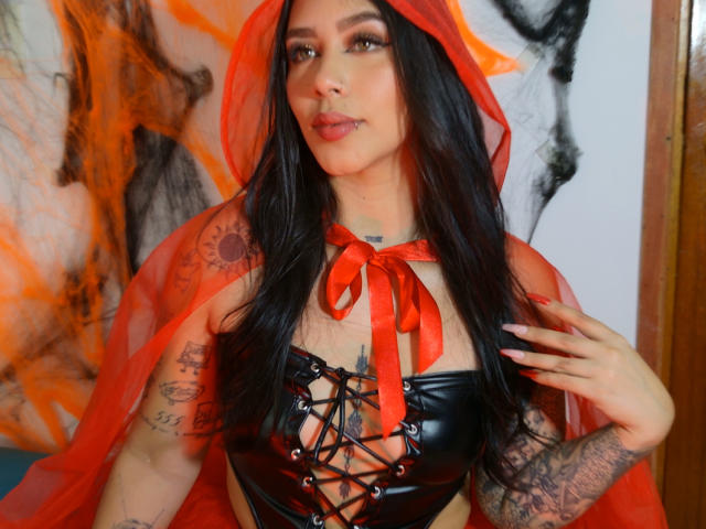 AgathaVelez69 with long black hair, red hood, and sheer red cape, wearing a black lace-up bustier, exposing her tits, tattoos on arms, and a red ribbon.
