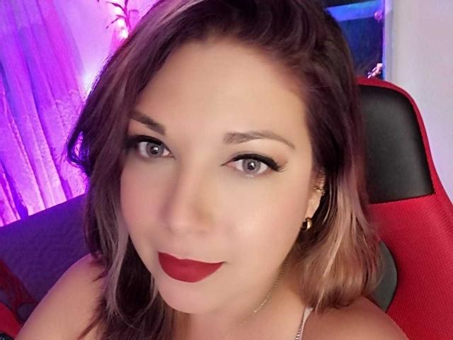 ScarletteThoms has red lipstick, black eyeliner, and dark brown hair with blonde highlights, sitting in a red chair, looking at the camera with a slight smile.