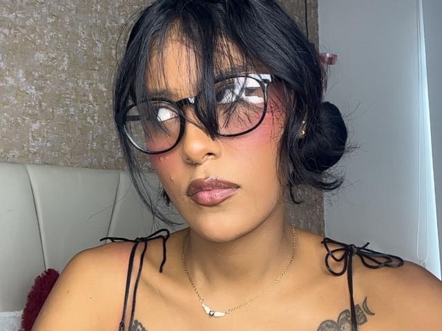 Babilett ha i capelli scuri, porta gli occhiali, ha un leggero rossore sulle guance e labbra rosa lucide. Indossa un top nero con spalline e piccoli tatuaggi sul petto. Si vede una collana..