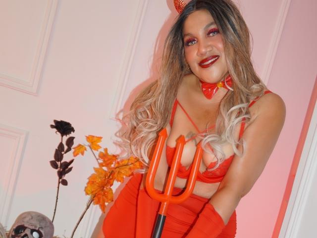 Mia Mancini mit langen blonden Haaren, roten Teufelhörnern und Make-up, trägt ein rotes Crop-Top, Handschuhe und einen Dreizack, lächelt in die Kamera und zeigt ihre großen Brüste..