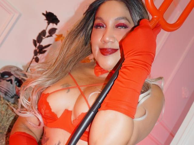 Mia Mancini mit rotem Lippenstift, roter Unterwäsche, roten Handschuhen, die einen roten Teufel-Dreizack hält, lächelnd, blondes Haar, große Brüste, Tattoos, rosafarbenes Zimmer..