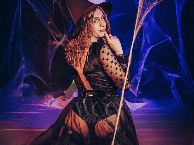AndyDupont in a witch outfit, black star-patterned sheer top, brown hat, curly hair, looking back, finger on chin, black belt, sheer black skirt, blue and purple lighting.