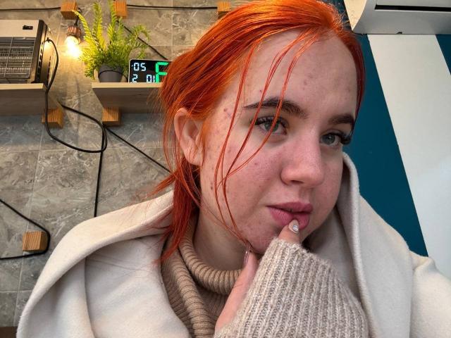 Casey Chase, con i capelli rossi, lentiggini e un piercing al naso, che morde un guanto beige in maglia senza dita, indossa un cappotto bianco e una collana di corda, e guarda la fotocamera..