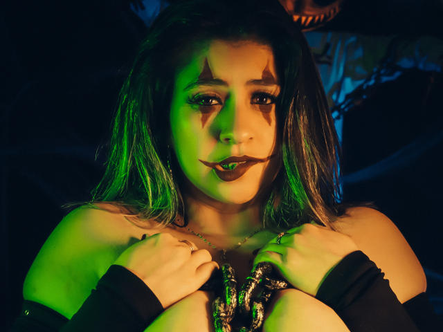 MelindaBoobs with green and blue lighting, dark makeup, and star-shaped face paint, holding a stringy, colorful necklace, wearing black sleeves, with her tits partially visible.