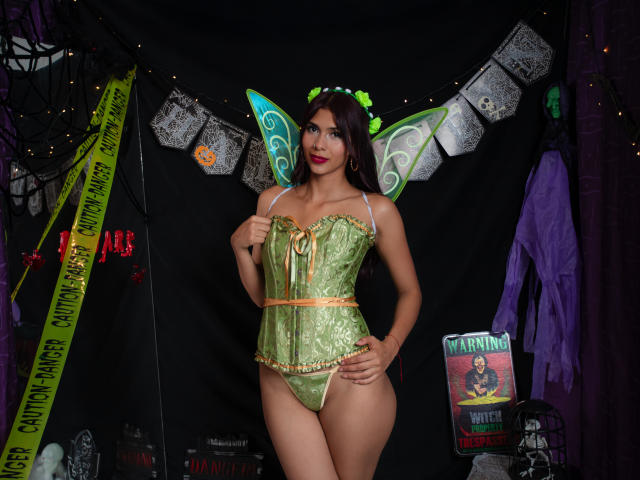 DanikaSerov with green fairy lingerie, green wings, and a leafy headband, standing with hands on hips, showing her tits and ass, in a dark, Halloween-themed room.