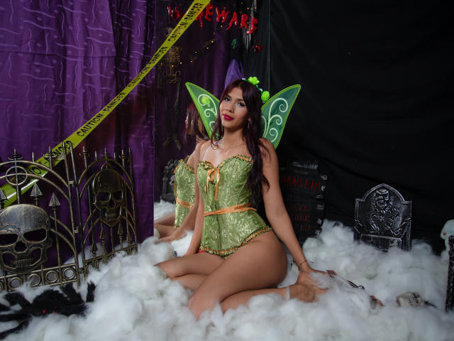 DanikaSerov, dressed in green fairy lingerie with butterfly wings, sitting on white fluffy ground, showing her tits and ass, with a skull and metal headpiece on the left.