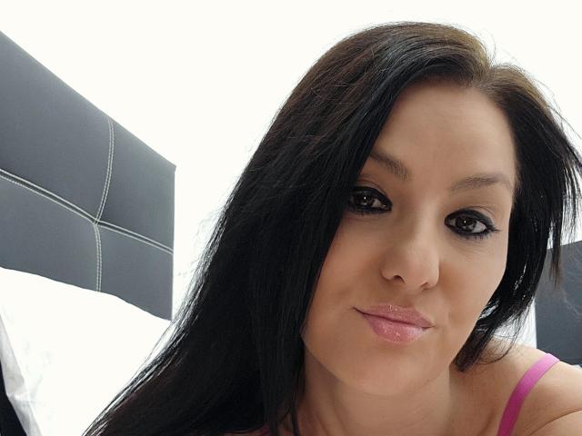 JoliDanielle has black hair, dark eyeliner, and pink lipstick, with a slight smile, wearing a pink top, showing her cute face and shiny lips. Her tits are small.