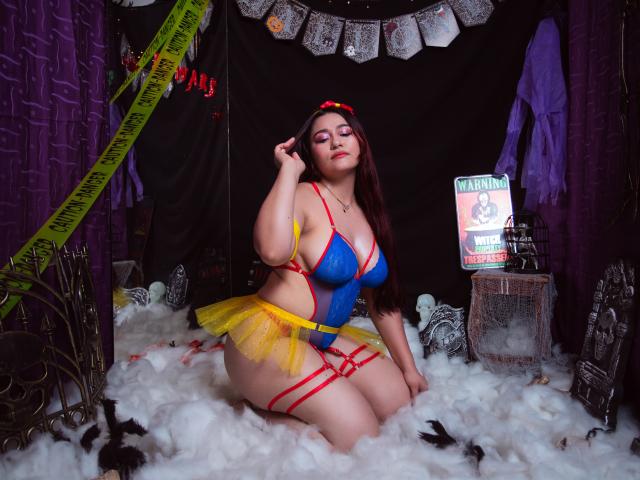 MaddieMoon con grandes senos, vestida con un disfraz de hada azul y amarillo, sentada sobre algo blanco y esponjoso, usando tirantes rojos, un lazo rojo y una falda amarilla, en una habitación de color púrpura oscuro..