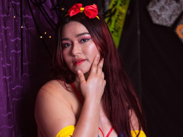 MaddieMoon with red lipstick, pink eyeshadow, and a red bow headband, touching her cheek, wearing a yellow top with red straps, and showing her small tits.