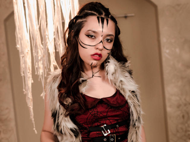 EllieCutee, com cabelo trançado, maquiagem preta no rosto, batom vermelho, vestindo uma blusa vermelha de renda, um cinto preto e um colete de pele, está em pé sob uma luz pendurada. Seios visíveis, com uma expressão séria..