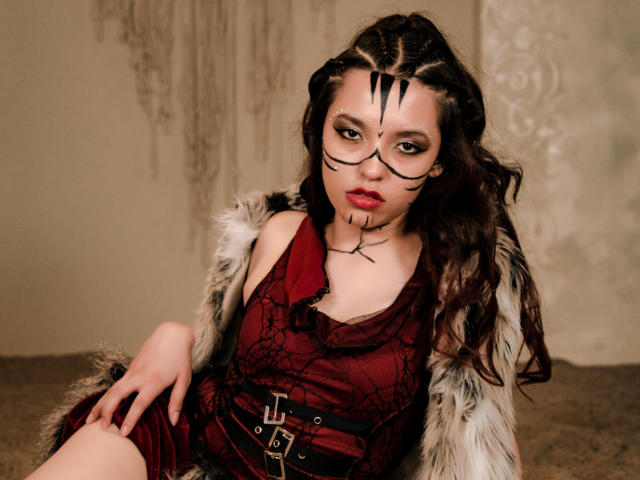 EllieCutee con trucco scuro sul viso, vestito rosso, cintura nera, cappotto di pelliccia, capelli castani ondulati, rossetto rosso, seduta, che guarda la fotocamera, espressione seria, sexy, pelle chiara..