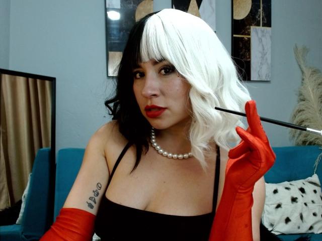 SaraKleine with black and white curly hair, red lipstick, black strap dress, red gloves, pearl necklace, holding a cigarette, tattoo on left arm, medium tits, looking at camera.