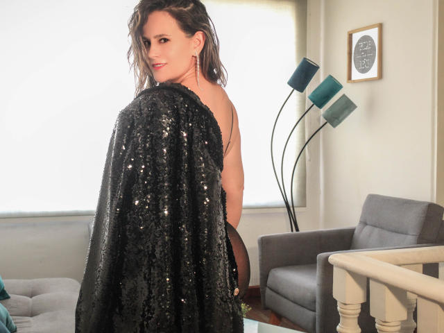 JeniElfie with wavy brown hair, a sequined black cape, and a sly smile, looking back over her shoulder, topless with visible tits, in a bright room.