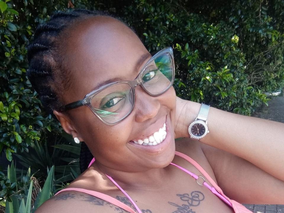 TheaMona with dark skin, braided hair, wearing glasses, pink top, smiling, showing white teeth, tattoo on chest, silver watch, and pearl earrings. Her ass is not visible.