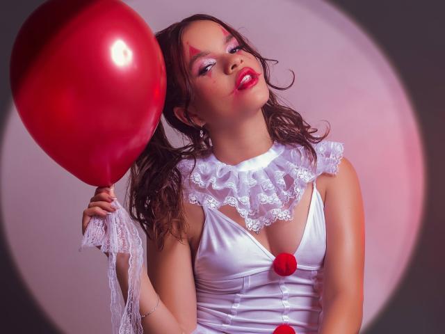 EmmaMarote with glossy red balloon, wearing white lace dress, red flower, and pink makeup, holding balloon to her right, winking with red lipstick, curly brown hair.