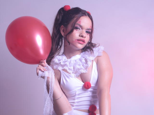 EmmaMarote with red balloon, white lace dress, and puffy red pom-poms, holding balloon to her right, pouting with pink face paint, dark hair in pigtails with red balls.