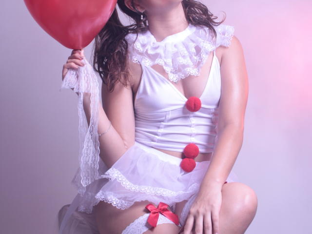 EmmaMarote with white lace outfit, red pom-poms, and white thigh-highs, holding a red balloon, kneeling, showing her tits and pussy, in a soft pink-lit photo.