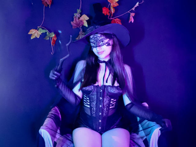 IrisBlair with long brown hair, black lace mask, top hat, black corset, gloves, holding a cigarette, sitting on a chair, purple blue lighting, leaves in background.