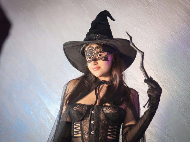 IrisBlair with witch costume, black lace mask, hat, holding stick, sheer gloves, lace-up corset, showing her tits, brown hair, white background, smiling.