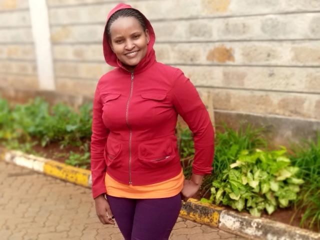 Emphi69 stands in front of a brick wall, wearing a red hooded jacket over an orange shirt and purple pants. She has a dark skin tone, braided hair, and a slight smile. The jacket has pockets and a zipper. She leans on a yellow-edged brick sidewalk with green plants in a small garden bed behind her. The image has a natural, casual feel.