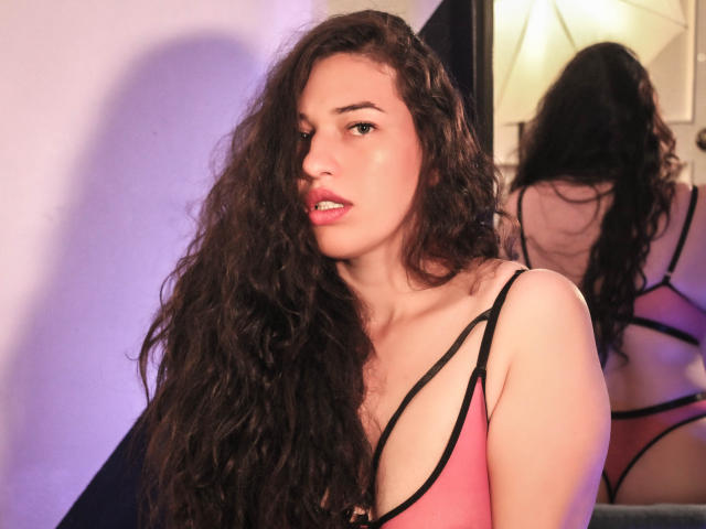 Rachel Wilson, con cabello castaño rizado y largo, usa un top delgado rosa y negro que deja ver sus senos. Está mirando a la cámara con los labios rosados ligeramente entreabiertos, y su reflejo se ve en un espejo. La iluminación es suave y morada..