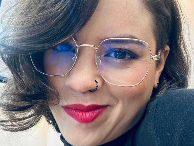 TaylorSexiest has blue eyes, wavy brown hair, a nose ring, and is wearing red lipstick with metal-framed glasses, looking slightly to the side with a subtle smile.