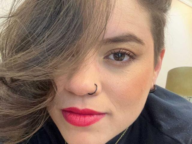 TaylorSexiest has a nose ring, red lipstick, and brown hair covering one eye. She's wearing a black shirt with a gold necklace. Her eyebrows are neatly shaped.