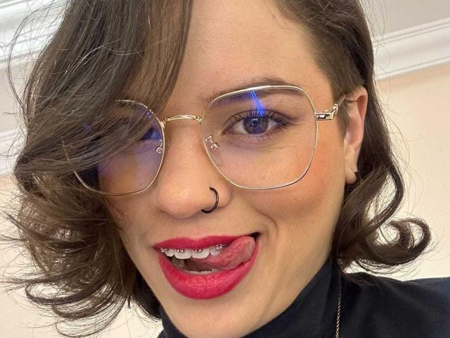 TaylorSexiest has wavy brown hair, gold glasses, a nose ring, red lipstick, and is sticking her tongue out. Her blue eyes are reflecting light.
