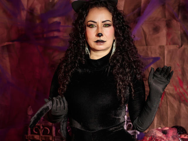 SamanthaAntolin stands in the center, wearing a black velvet dress and long black gloves. She has curly dark hair, green eye makeup, and dark lipstick. She holds a black cat tail in her right hand and has a black nose dot on her face. The background is a brown, textured wall with purple and red spray paint. She wears a black cat ear headband and has silver chandelier earrings. The image is a photograph with a dramatic, goth aesthetic.