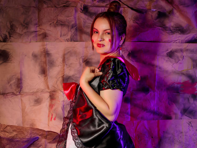 VictoriiaJones com maquiagem vermelha no rosto, vestindo um vestido preto e vermelho, em pé com as mãos perto do peito, cabelo castanho preso em um coque, iluminação roxa e rosa, sorriso confiante..