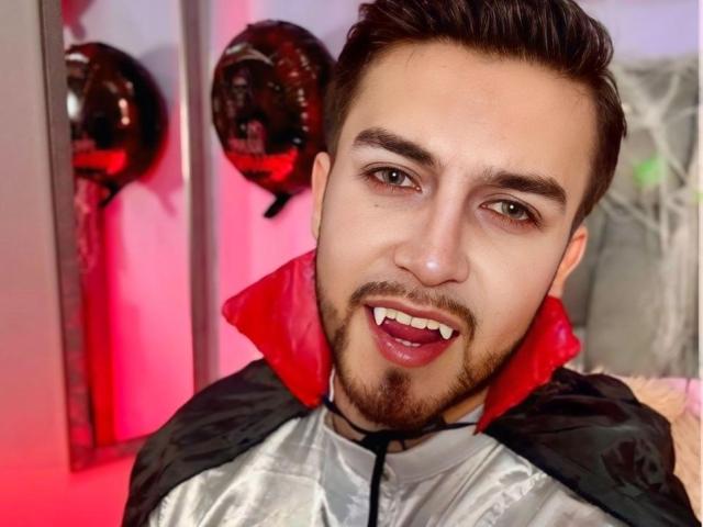 DaltonWhite vestito da vampiro, con un mantello nero e rosso, trucco con canini, e capelli castani corti, sorride con le labbra leggermente dischiuse, in una stanza luminosa con illuminazione rossa..